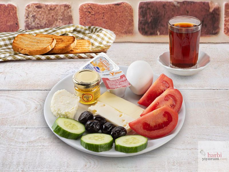 Bir kahvaltı tabağı görseli: Kızarmış ekmek dilimleri, bir bardak çay, haşlanmış yumurta, bal, tereyağı, siyah zeytin, taze salatalık dilimleri, dilimlenmiş domates, beyaz peynir ve kaşar peynirinden oluşan sade bir kahvaltı sunumu.