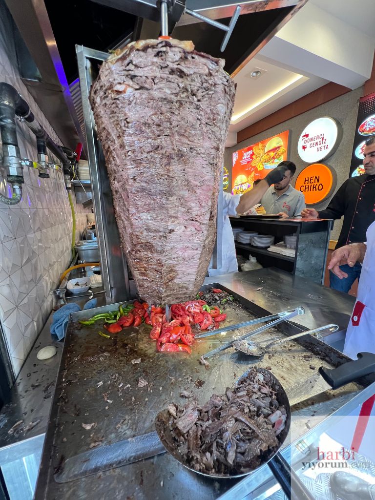 Cengiz Usta'nın Beşiktaş'taki yeni döner büfesinde döner tezgahı. Büyük bir et şişi döner kesim için hazır, önünde ise taze domates ve biberler bulunuyor. Dönerin alt kısmında kesilmiş döner parçaları, servis edilmek üzere tezgah üzerinde yer almakta. Arka planda 'Dönerci Cengiz Usta' tabelası ve Henchiko Fried Chicken logosu görülebiliyor.