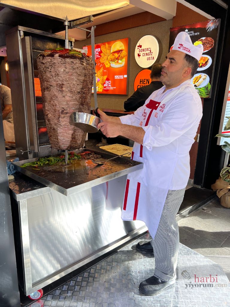 Cengiz Usta, Beşiktaş'taki yeni döner büfesinde döner tezgahının başında döneri keserken. Klasik beyaz şefi önlüğü ve tabelada 'Dönerci Cengiz Usta' yazısıyla, usta döner hazırlığı yapıyor. Arka planda Henchiko Fried Chicken tabelaları görülebiliyor, bu da iki işletmenin aynı alanda faaliyet gösterdiğini işaret ediyor.