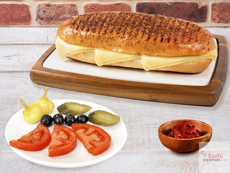 Çift kaşarlı tost görseli: Susamlı çıtır ekmek arasında eritilmiş bol kaşar peyniriyle hazırlanan tost, yanında domates dilimleri, siyah zeytin, kornişon turşu, acı biber turşusu ve bir kase acı ezme ile sunulmuş şık bir kahvaltı tabağı.