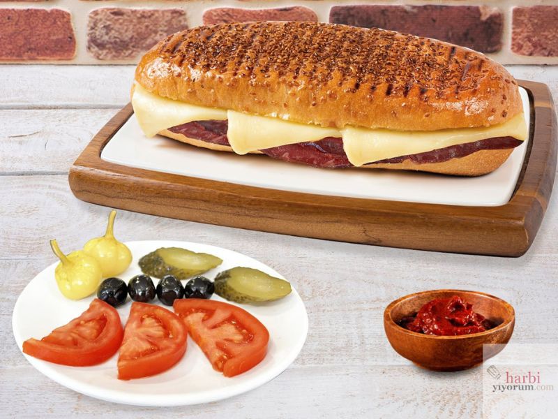 Bir pastırmalı kaşarlı tost görseli: Üzeri susamla kaplı çıtır bir ekmek arasında pastırma ve eritilmiş kaşar peyniri, yanında domates dilimleri, kornişon turşu, siyah zeytin, acı biber turşusu ve bir kase acı ezme ile sunulmuş lezzetli bir kahvaltı tabağı.