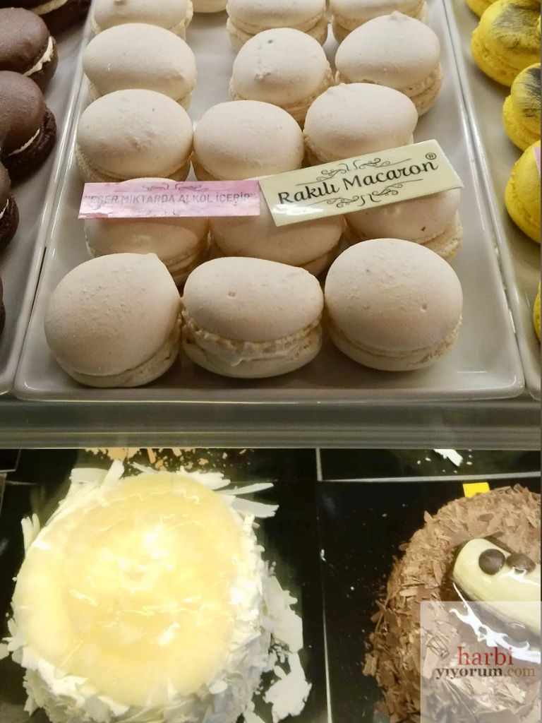 A La Creme pastanesinde sergilenen tatlılar. Üst rafta beyaz renkli "Rakılı Macaron" etiketli macaronlar, yanlarında ise "Eser miktarda alkol içerir" uyarısı yer alıyor. Alt bölümde ise beyaz çikolata kaplamalı bir pasta ve çikolata parçalarıyla süslenmiş başka bir tatlı görülmekte. Görsel, mekanın farklı ve özgün tatlılarını vurguluyor.