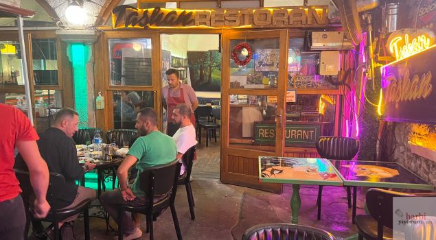 Taşhan Restoran Bartın'ın dış cephesi, açık alanda oturan misafirler ve neon ışıklarla süslenmiş meyhane giriş kapısı. Taşhan Meyhane'nin otantik atmosferini yansıtan bir akşam görüntüsü.